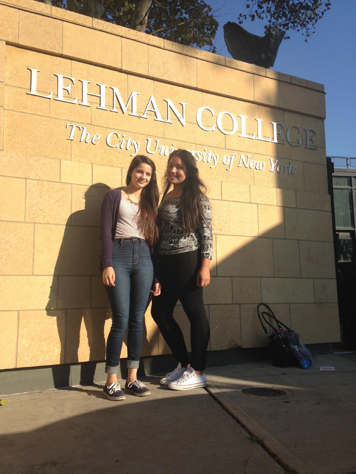 Andrea and Yessenia standing in front of an engraving at Lehman College featuring the school's name and slogan
